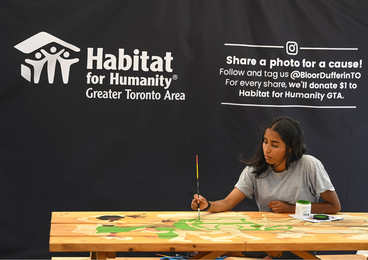 A person painting artwork on a wooden table in front of a Habitat for Humanity GTA promotional display.