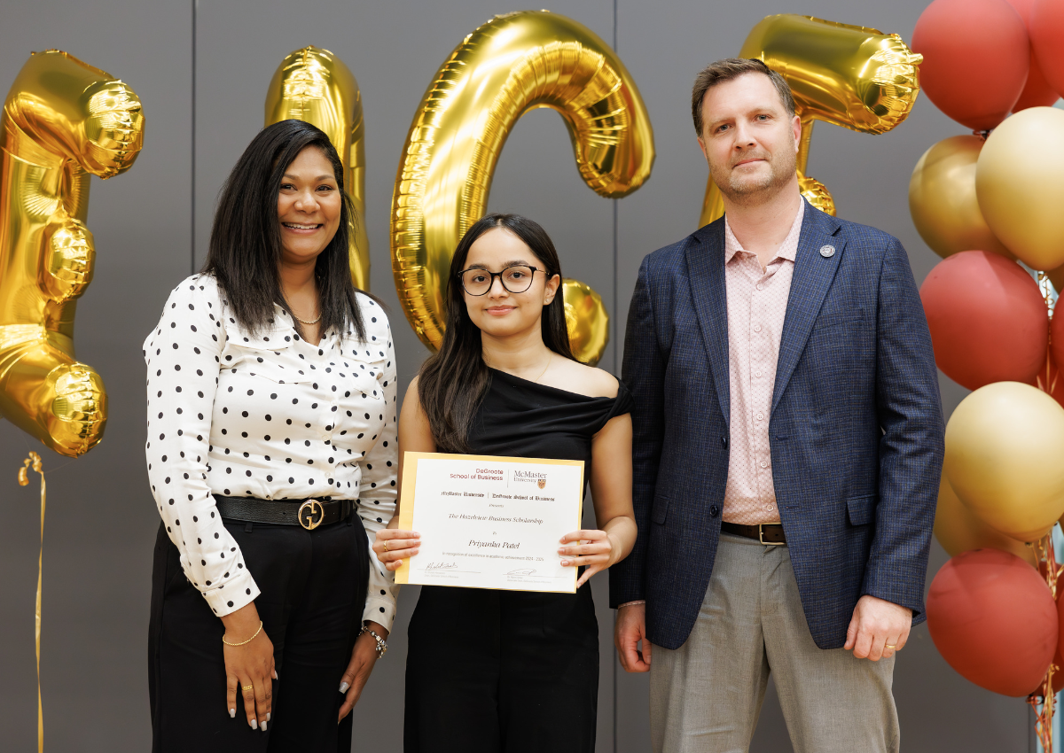 Three people standing together indoors, one holding a certificate, with gold number balloons in the background.