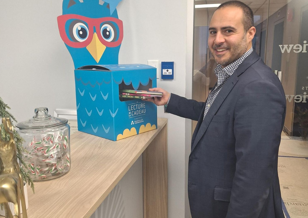 A person places a book into a blue donation box shaped like an owl on a countertop.